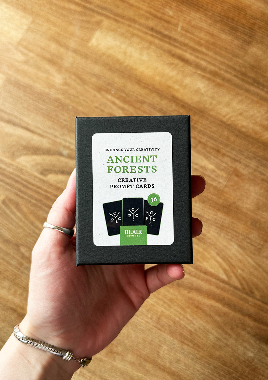 Hand holding a box of 'Ancient Forests' creative prompt cards on a wooden surface