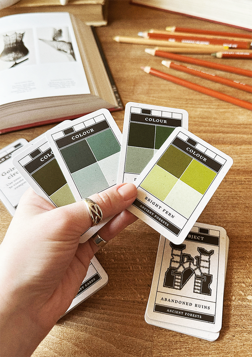 Color swatch cards held by a hand on a wooden surface with pencils and books in the background.