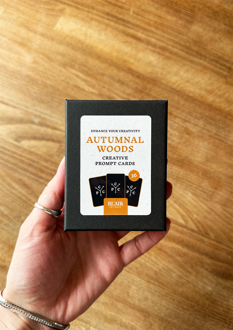 Hand holding a box of 'Autumnal Woods' creative prompt cards on a wooden surface