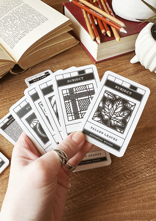 Hand holding patterned cards on a wooden surface with books and pencils in the background