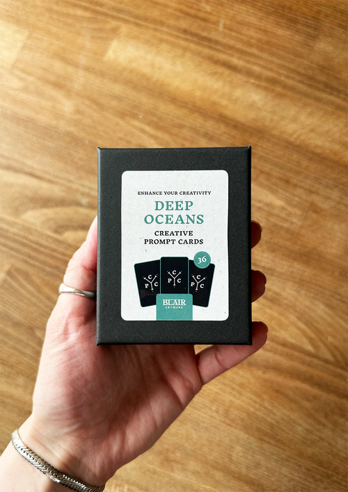 Hand holding a box of 'Deep Oceans' creative prompt cards on a wooden surface