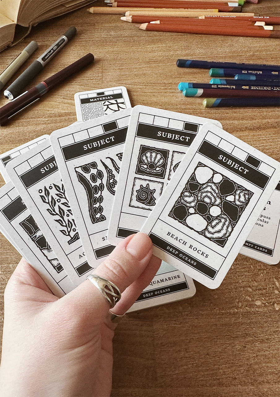 Hand holding black and white cards with nature-themed designs on a wooden surface with art supplies.