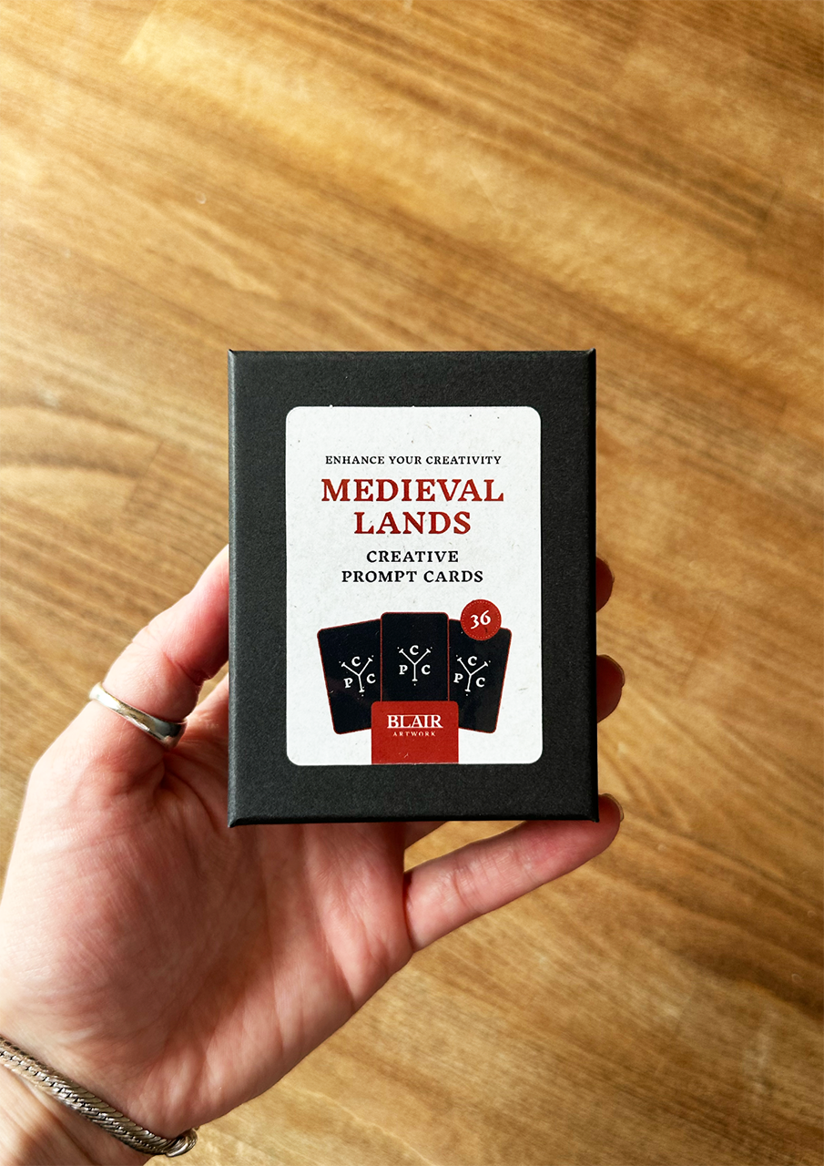 Hand holding a box of 'Medieval Lands' creative prompt cards on a wooden surface