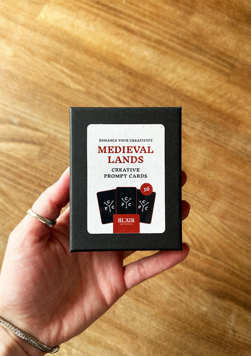 Hand holding a box of 'Medieval Lands' creative prompt cards on a wooden surface