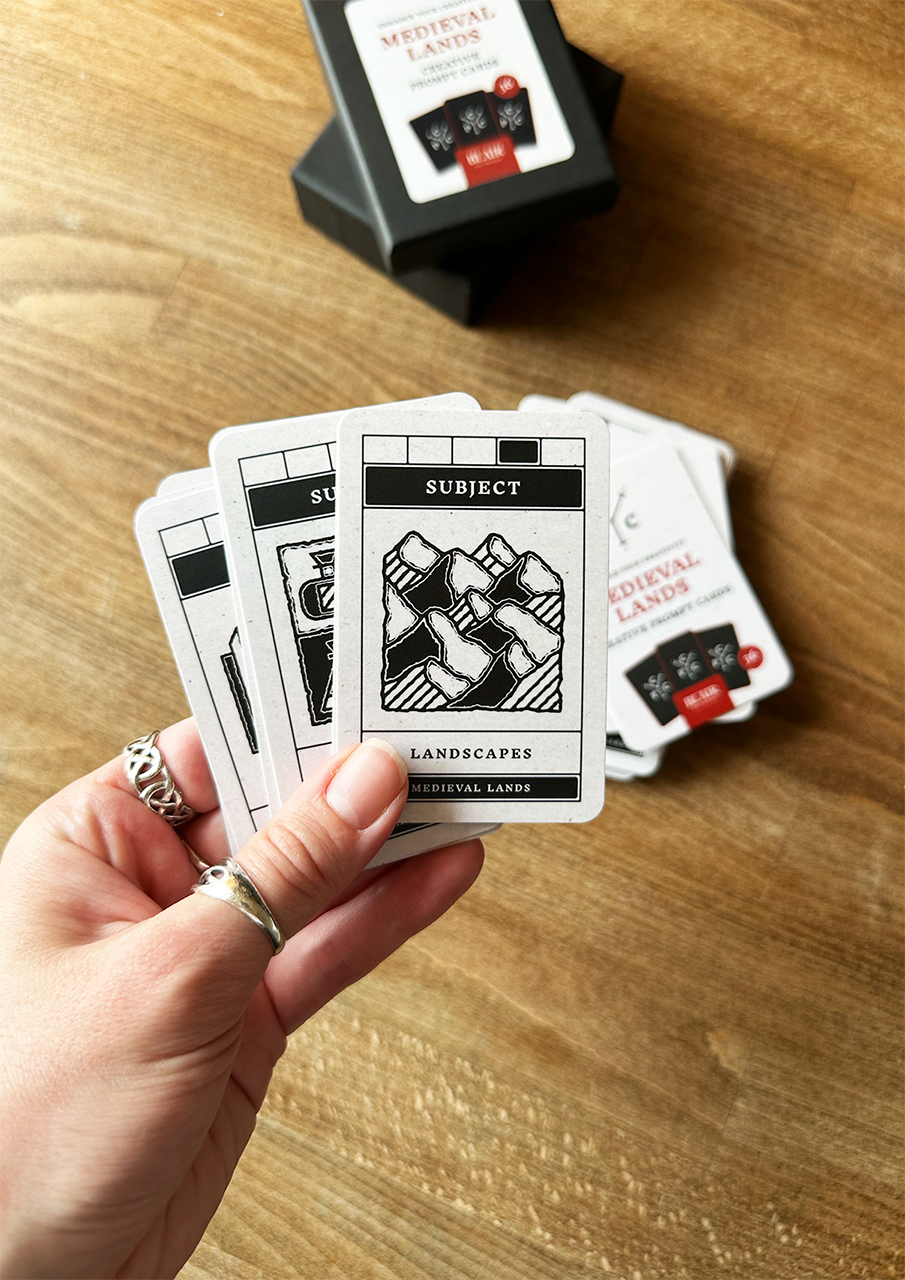 Hand holding playing cards with 'Medieval Landscapes' theme on a wooden surface