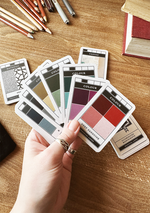 Color swatch cards held by a hand on a wooden surface with colored pencils and books.