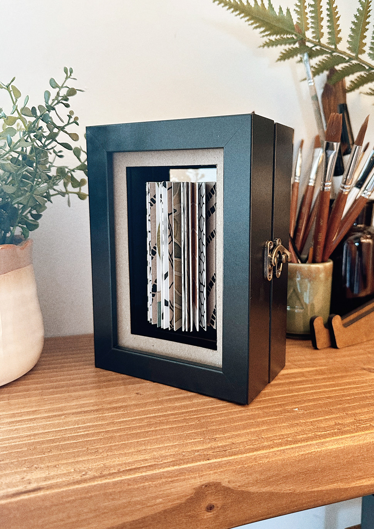 Artwork in box on shelf with paintbrushes and plants