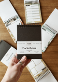 A hand holding a compact black pocketbook planner with watercolour backgrounds, surrounded by other similar planners laid out on a wooden surface.