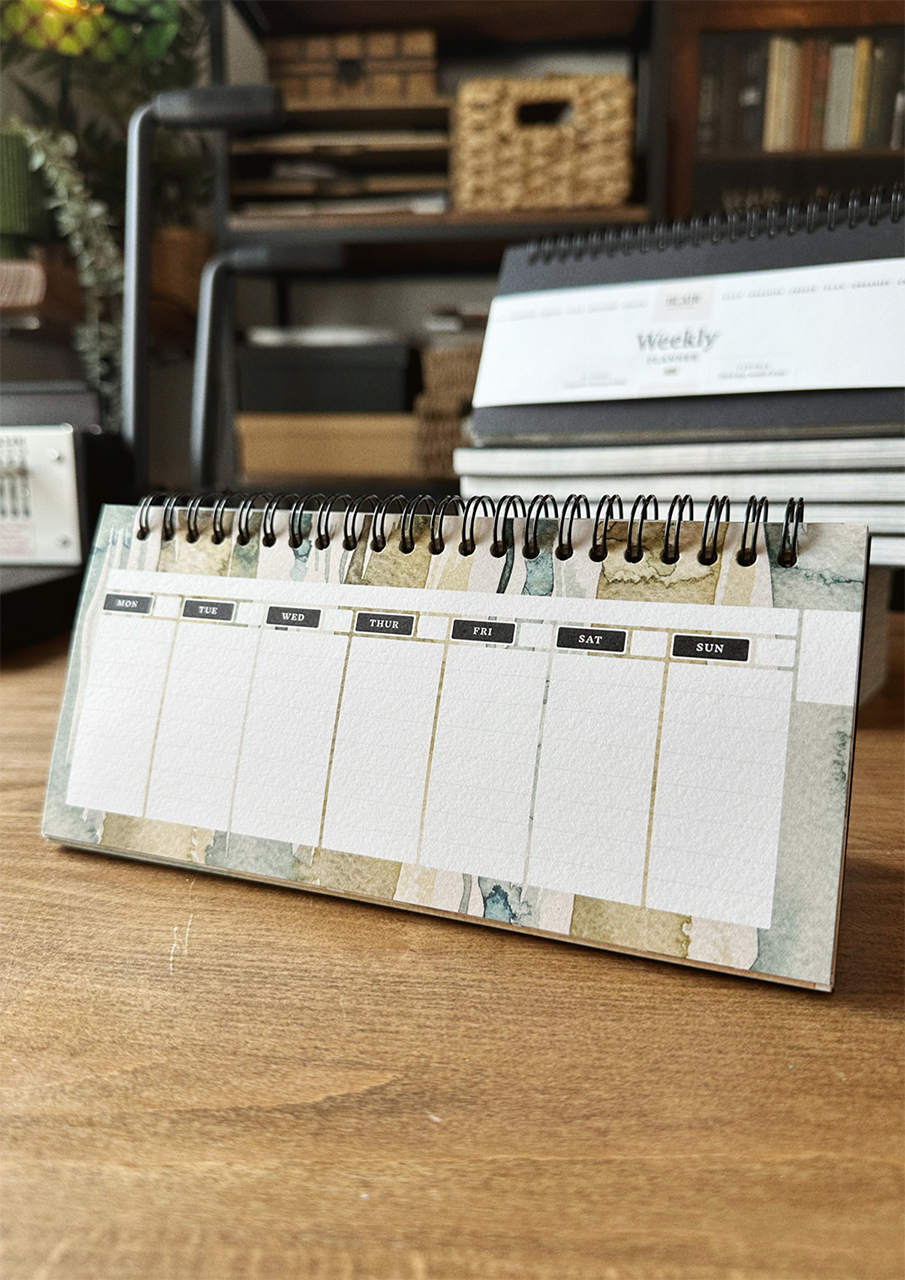 A desktop weekly planner with a watercolor design, displayed on a wooden surface.