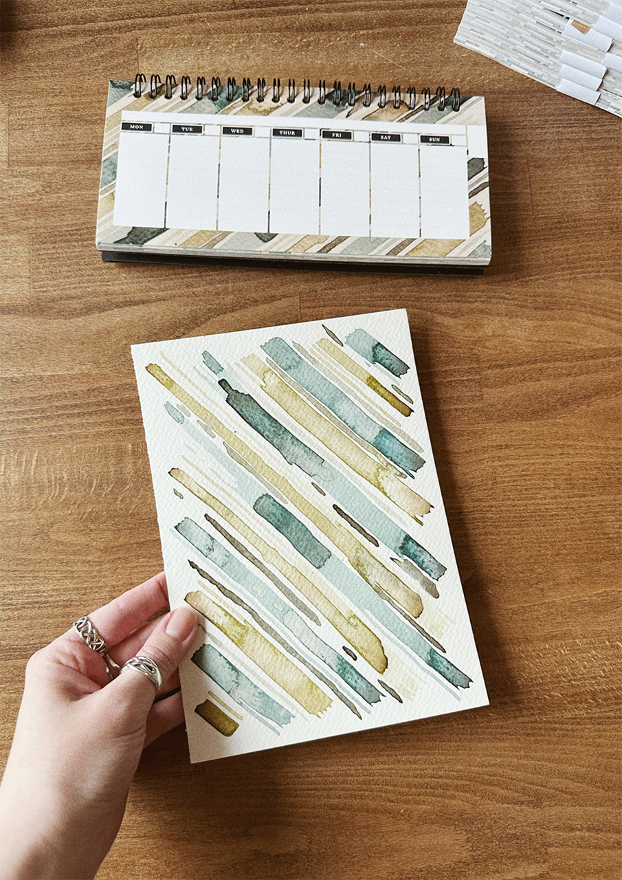 A desktop weekly planner, displayed on a wooden surface with watercolour painting