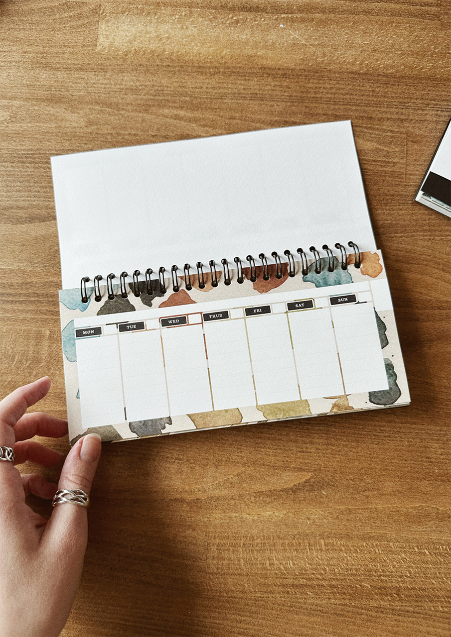 A desktop weekly planner with a watercolor design, displayed on a wooden surface.