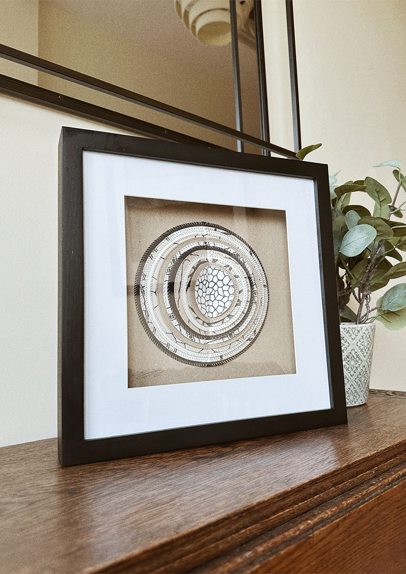 A framed hand-stitched artwork depicting a large circular paper moon in a simple black frame, set on a shelf with a plant and mirror