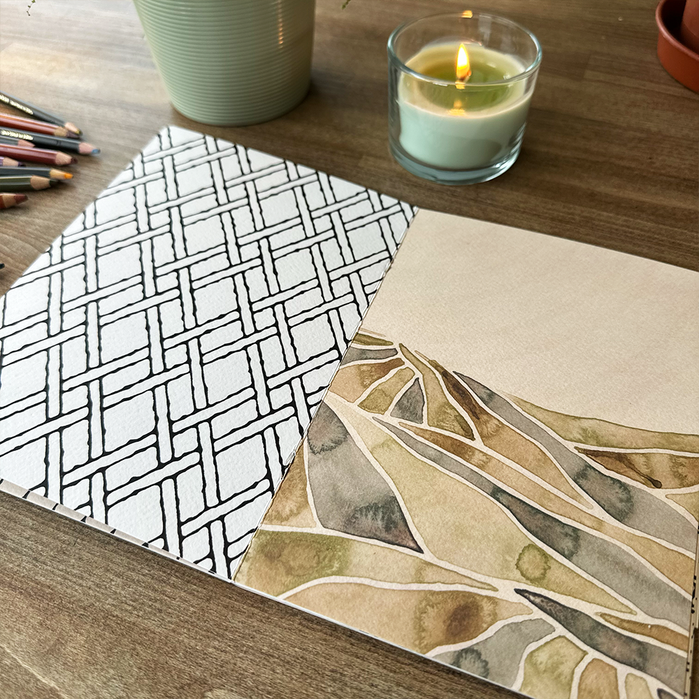 Two pages with different patterns on a wooden surface with a candle and pencils.