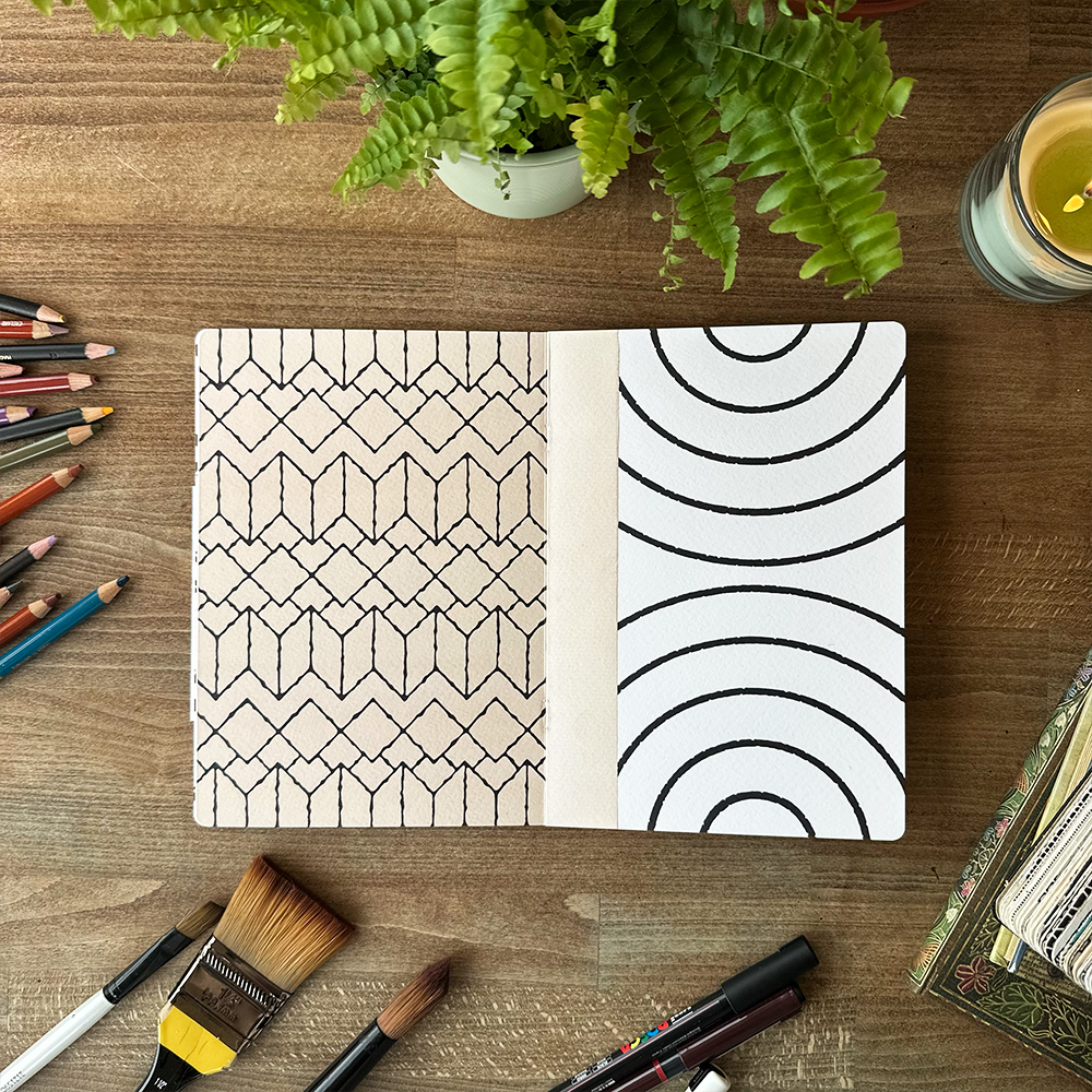 Notebook with geometric patterns on a wooden surface with art supplies and a plant.