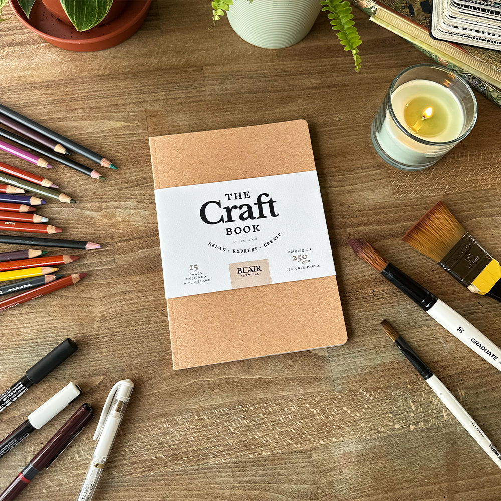 Kraft paper notebook labeled 'The Craft Book' surrounded by art supplies on a wooden surface.