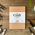 Brown notebook titled 'The Craft Book' on a wooden surface with art supplies.