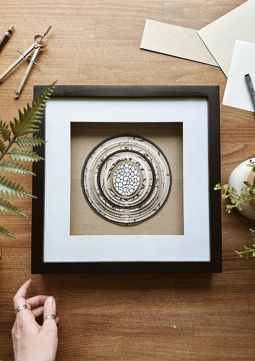 A framed hand-stitched artwork depicting a large circular paper moon in a simple black frame, set against a wooden table and decorative background.
