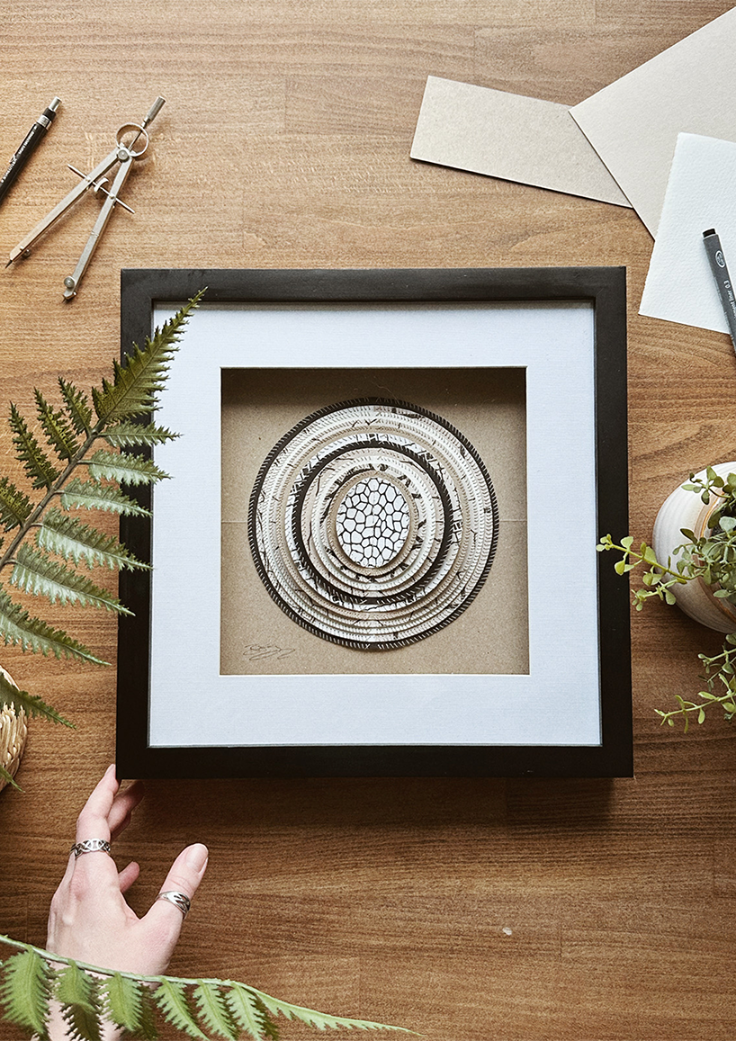 A framed hand-stitched artwork depicting a large circular paper moon in a simple black frame, set against a wooden table and decorative background.