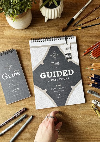 Two Guided Illustration books on display on a wooden desk surrounded by pens, pencils, plants and a hand