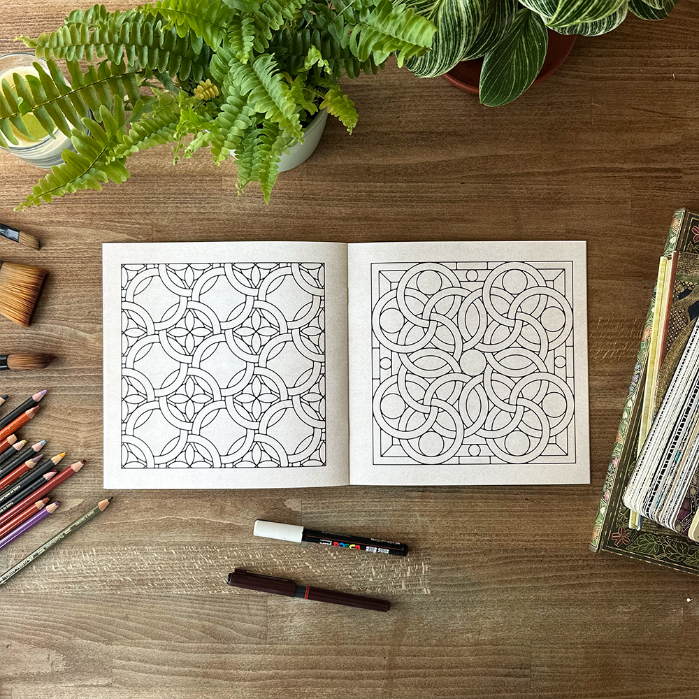 Open coloring book with intricate patterns on a wooden table with art supplies and plants.