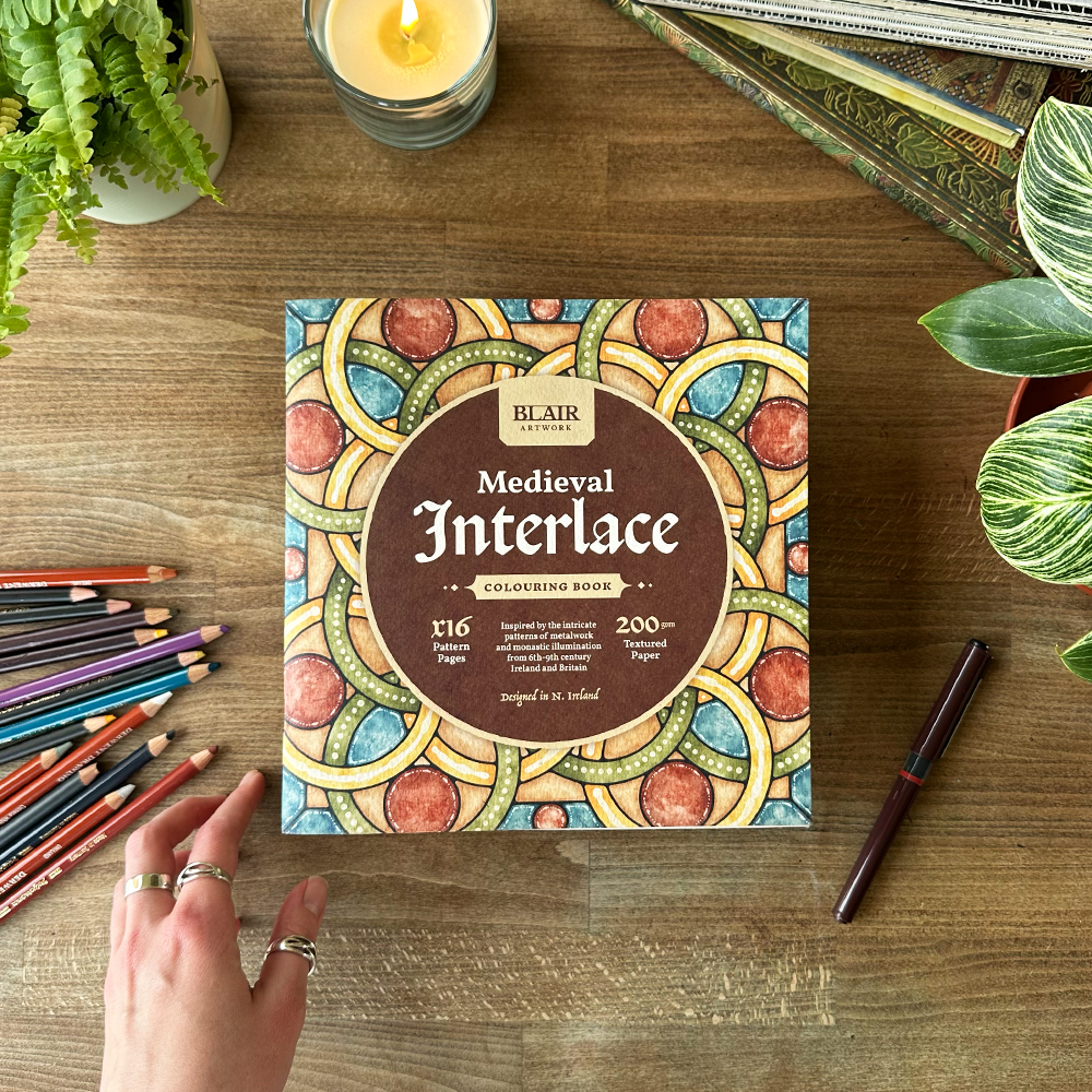 Blair Coloring Book titled 'Medieval Interlace' on a wooden table with coloring pencils and a candle.