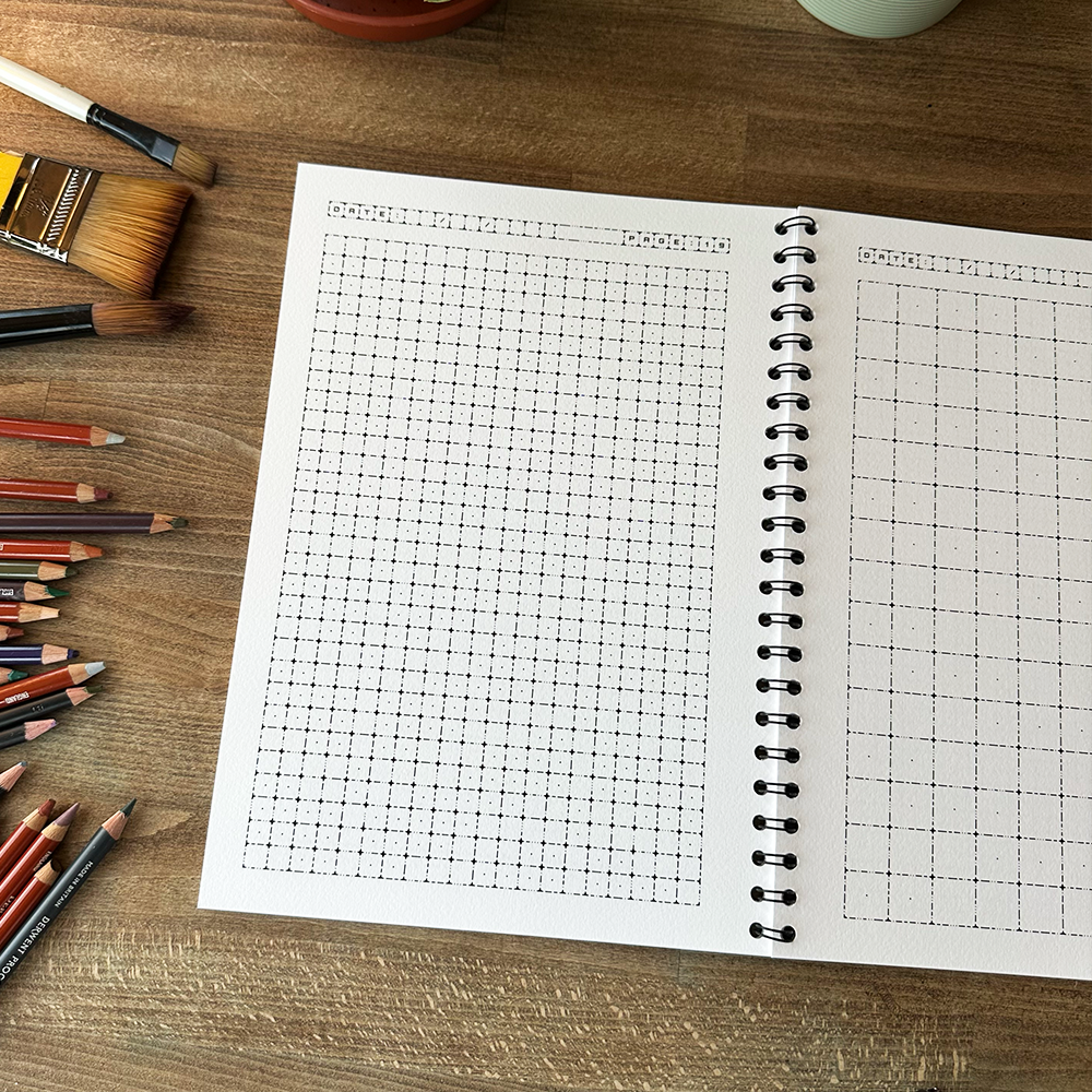 Open notebook with grid paper on a wooden surface with pencils and paintbrushes.