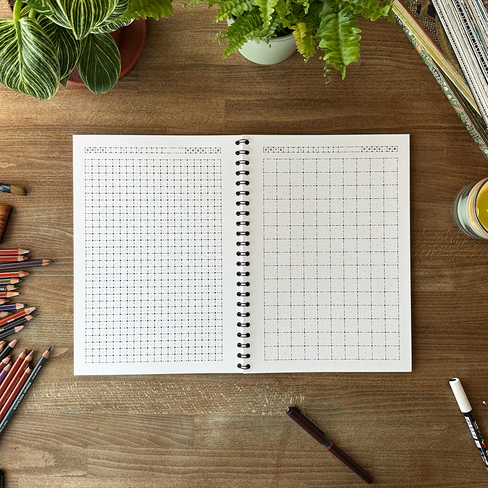 Open spiral-bound notebook with grid paper on a wooden desk with stationery items and plants.