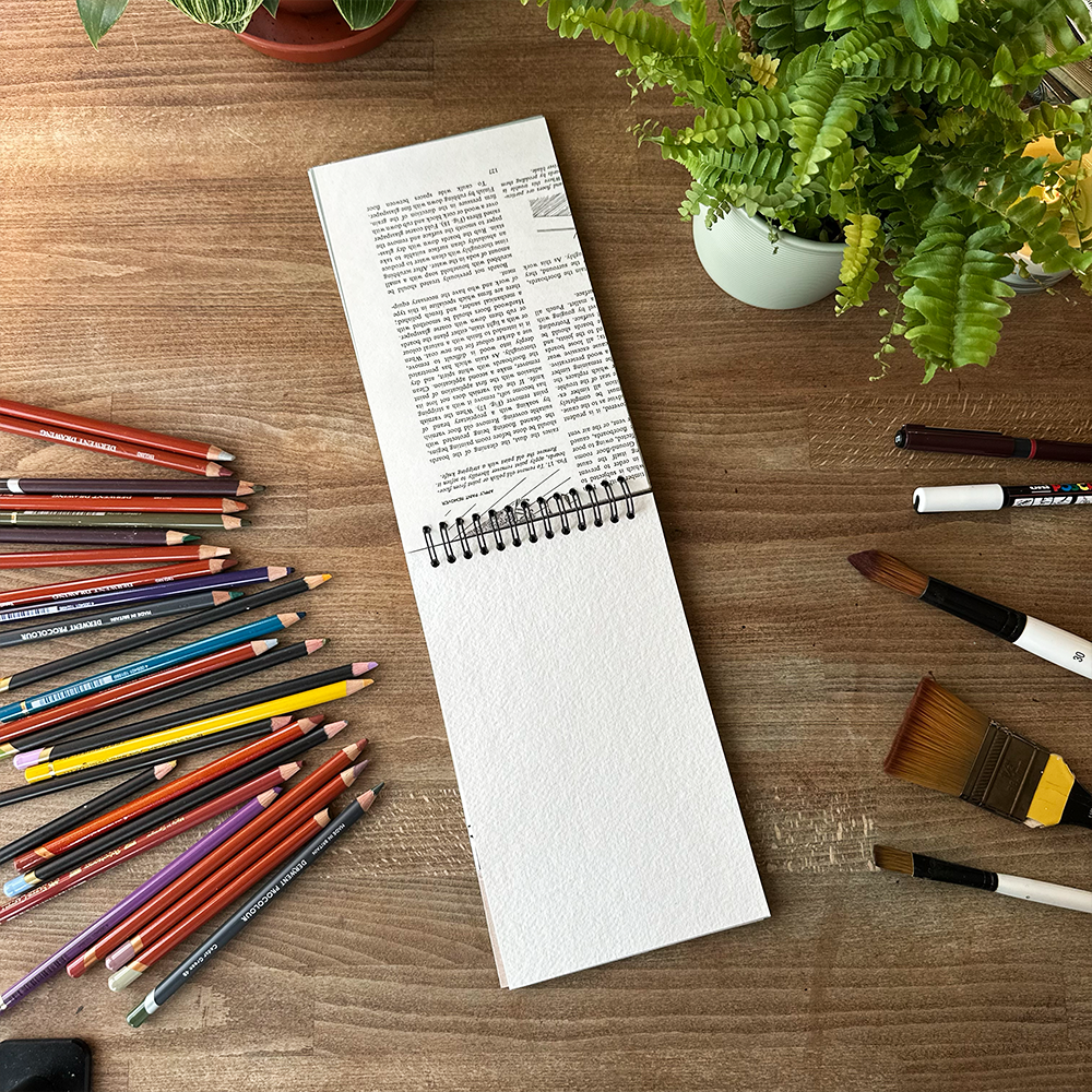 Notebook on a wooden surface with colored pencils, markers, and plants.