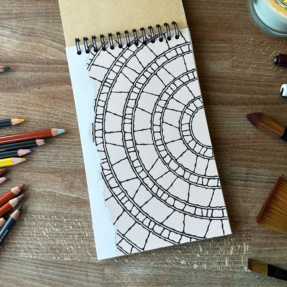 Notebook with a black and white geometric pattern on a wooden surface with art supplies.