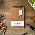 Brown notebook with 'The Scrap Book' tag on a wooden surface with art supplies.