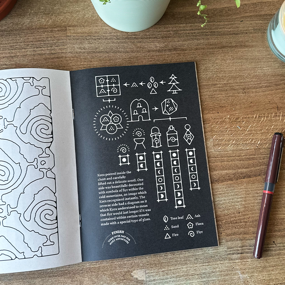 Open book with black and white illustrations on a wooden surface with a pen and small plant.