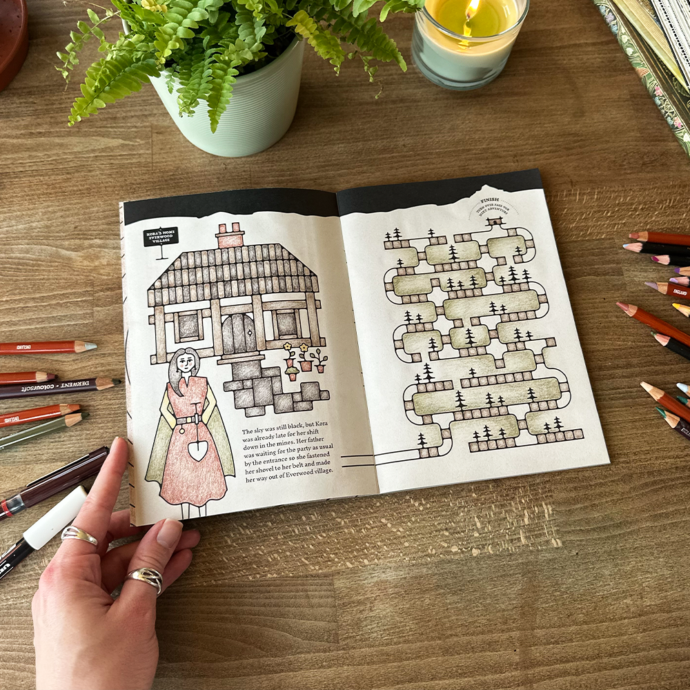 Open book with illustrations on a wooden table with pencils and a candle.