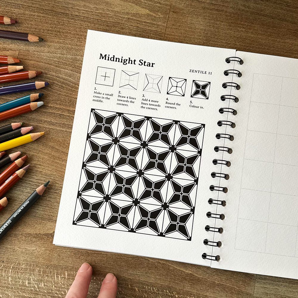 Open notebook with geometric design and coloring instructions on a wooden surface with colored pencils.